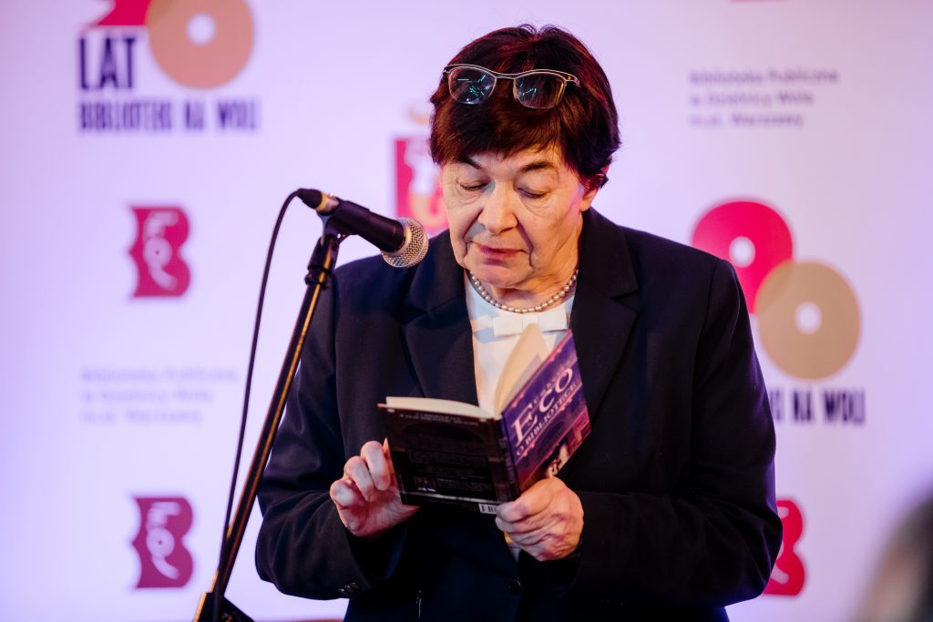 Kobieta przemawia przez mikrofon. W rękach trzyma książkę Umberto Eco.